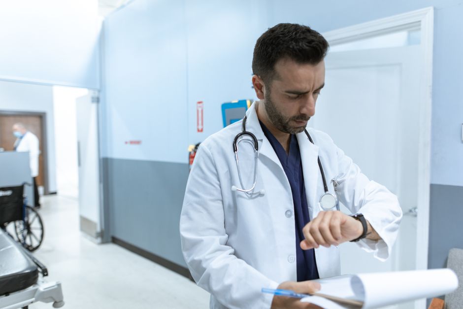 Médico em ambiente hospitalar verificando o horário em seu smartwatch, simbolizando o cuidado com o tempo e a atenção ao paciente.