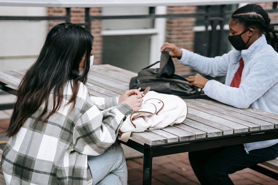 Duas jovens conversando ao ar livre, usando máscaras e compartilhando momentos de convivência com dispositivos móveis e mochilas ao redor.