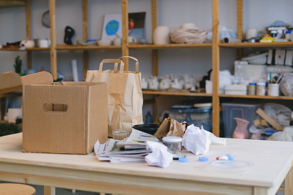 Imagem de espaço de trabalho desorganizado com caixas, embalagens, papéis amassados e materiais diversos, sugerindo um ambiente de produção artístico ou artesanal.