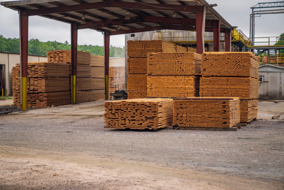 Pilhas de madeira empilhadas em uma área exterior de uma fábrica de materiais de construção, pronta para uso ou transporte.