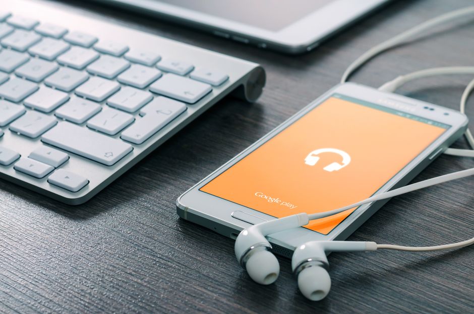 Imagem de um teclado, um smartphone com o aplicativo Google Play aberto e fones de ouvido, sugerindo uso de tecnologia para comunicação ou trabalho remoto.