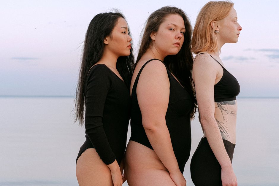 Três mulheres de diferentes tamanhos e aparências posando juntas à beira do mar ao pôr do sol, transmitindo uma mensagem de diversidade e autoestima.