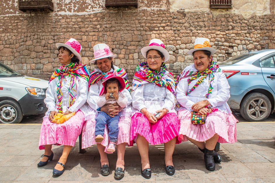 Mulheres idosas em vestimentas tradicionais coloridas, sentadas em uma praça em frente a uma parede de pedra, participando de uma celebração cultural ou festiva.