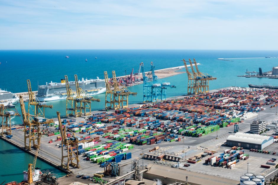 Imagem de um porto de carga com vários contêineres, guindastes e navios ao fundo, representando o tema de logística e comércio internacional.