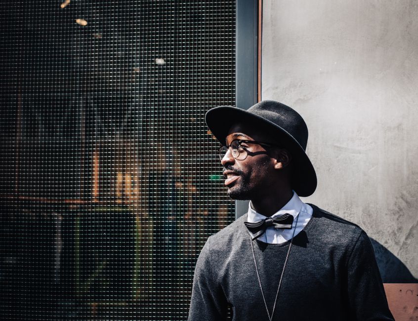Homem elegante com chapéu e óculos, posando ao lado de uma parede de vidro, transmitindo estilo e personalidade para um blog de moda ou estilo de vida.