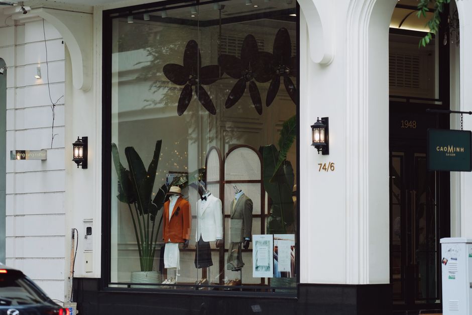 Vitrine de loja de roupas masculinas com manequins exibindo ternos e jaquetas, localizada em uma rua urbana.