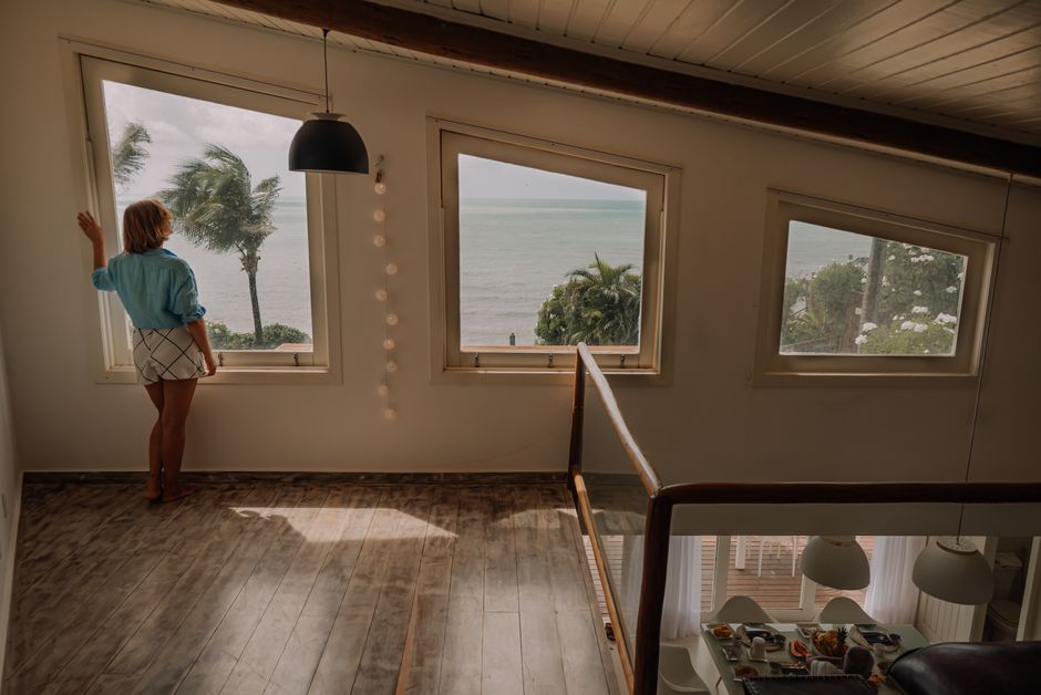 Mulher observando o mar em uma casa com janelas de vidro grande, criando uma atmosfera tranquila e contemplativa.