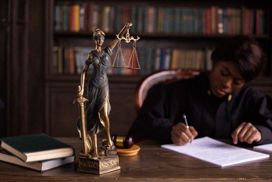 Imagem de uma advogada ou juíza estudando e tomando notas em um escritório com uma escultura da Justiça ao lado, simbolizando o tema jurídico e legalidade.