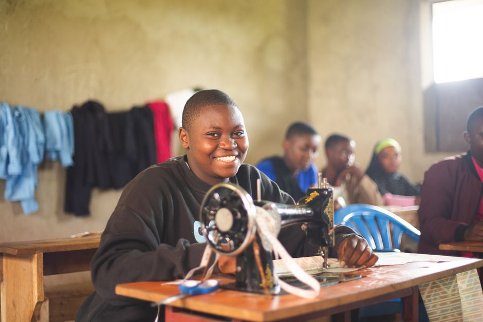 Jovem costurando com uma máquina de costura manual em um ambiente escolar, promovendo educação profissional para meninas na África.