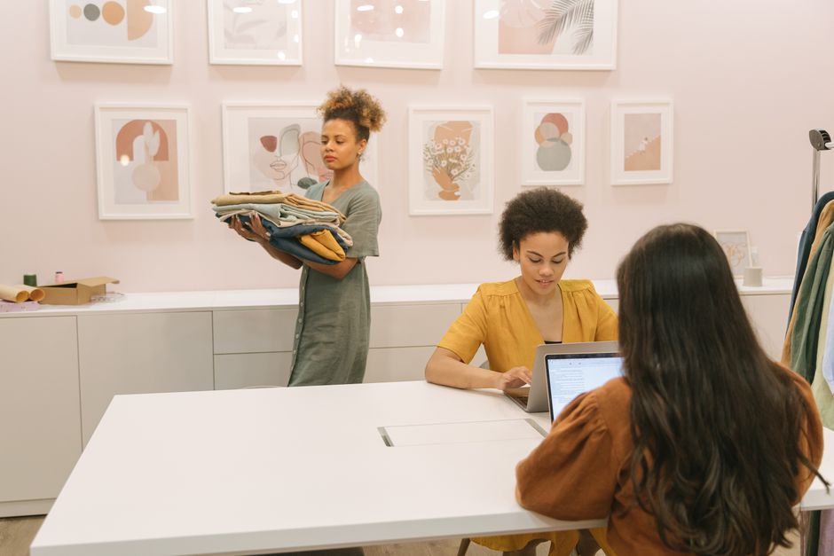 Imagem de colegas de trabalho em escritório, com uma delas organizando roupas enquanto as outras trabalham em computadores, sugerindo uma discussão sobre moda ou gestão de inventário.