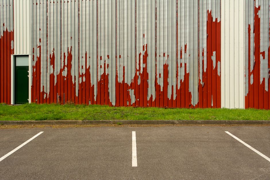 Imagem de uma parede de metal desgastada com pintura vermelha descascando, ao fundo uma área de estacionamento vazia.