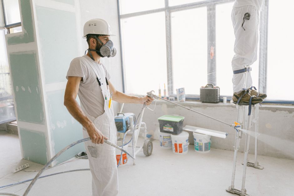 Profissional de construção usando máscara de gás e equipamento de proteção ao aplicar tinta ou impermeabilizante em uma parede interna de um edifício em reforma.