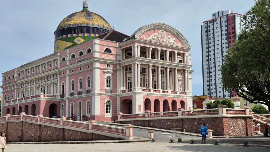 Imagem de um edifício histórico com arquitetura elegante, combinando elementos clássicos e modernos, situado em uma área urbana com alguns árvores e pessoas ao redor.