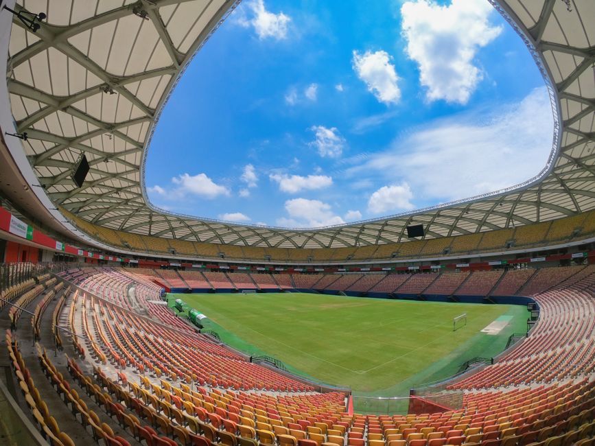 Visão interna de um estádio de futebol ao ar livre, mostrando o campo e as arquibancadas sob um céu parcialmente nublado.