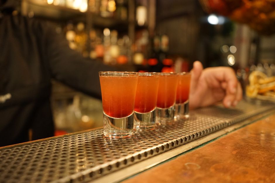 Imagem de quatro drinques de cor âmbar sobre uma bancada de bar, com um bartender ao fundo, em um ambiente acolhedor e iluminado.