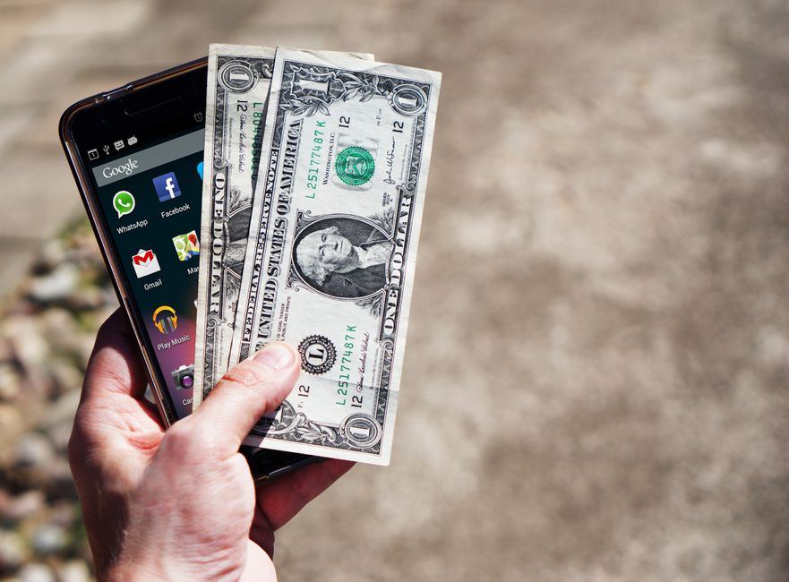 Imagem de uma mão segurando um smartphone com notas de dinheiro, simbolizando o tema de finanças digitais e pagamentos móveis.