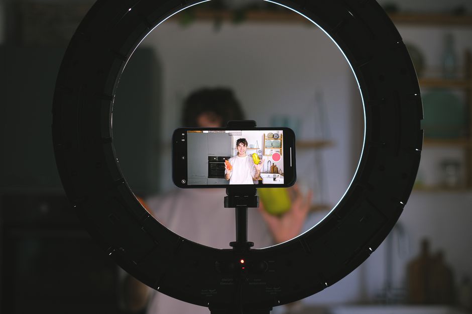 Pessoa gravando vídeo de culinária ou lifestyle usando um ring light para melhorar a iluminação, com foco na câmera que captura a pessoa ao fundo.