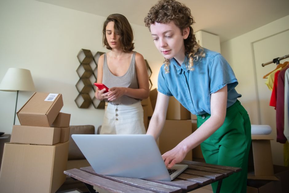 Duas mulheres organizando um espaço de trabalho com caixas e computador, sugerindo planejamento e organização de negócios ou mudança de loja virtual.