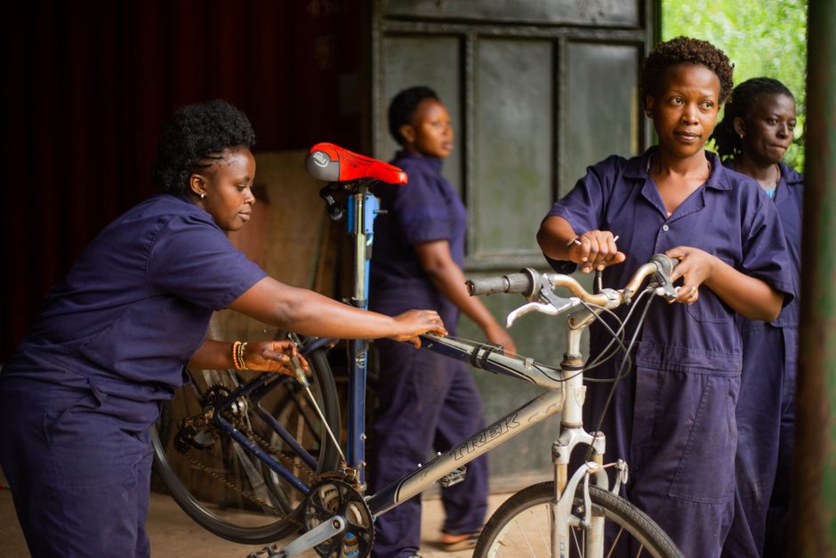 Mulheres vestidas com uniformes trabalhando na manutenção de bicicletas em um ambiente de oficina comunitária.