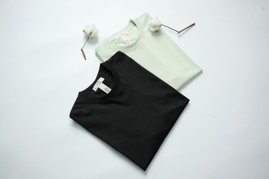 Duas camisetas básicas, uma preta e uma branca, dispostas lado a lado em um fundo branco minimalista, com detalhes de algodão em caixinha de fósforo.