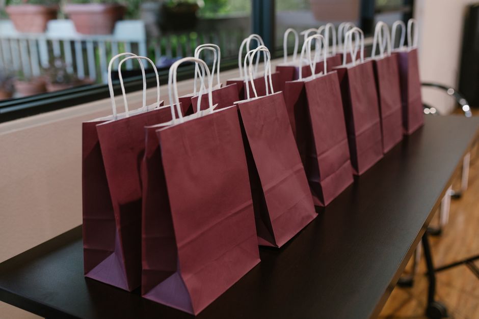 Imagem de várias sacolas de papel na cor vinho alinhadas sobre uma mesa ao lado de uma janela, transmitindo organização para uma ocasião especial.