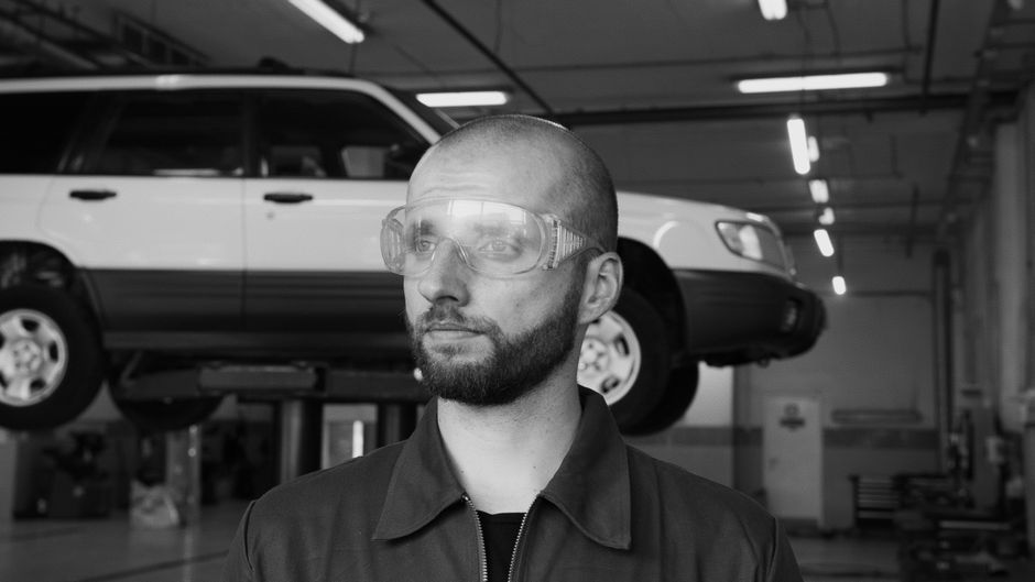 Homem em uma oficina mecânica, analisando um carro que está suspenso em um elevador para manutenção.