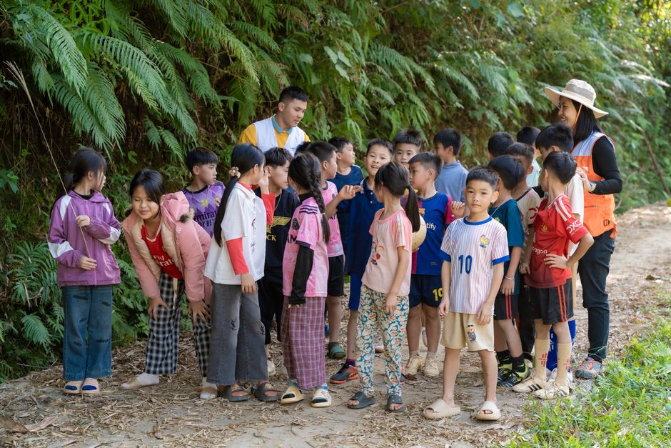 Grupo de crianças explorando a natureza com apoio de uma monitora durante uma atividade ao ar livre em um ambiente escolar ou recreativo.