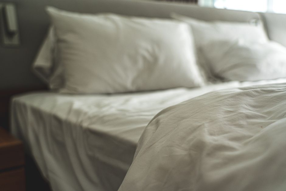 Cama desfeita com roupas de cama brancas em um quarto com iluminação natural, transmitindo uma sensação de conforto e rotina diária.