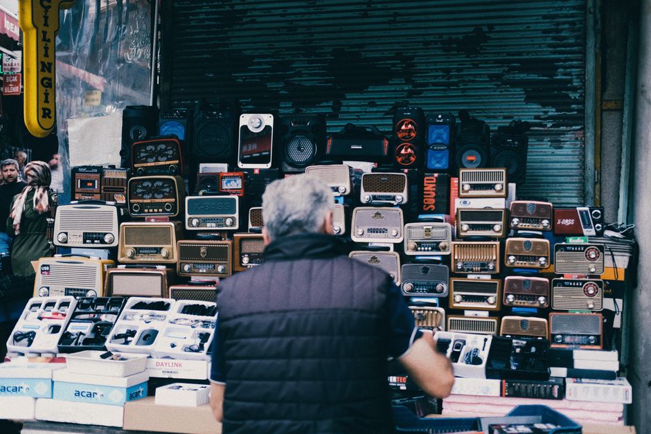 Homem observando uma banca de rádios vintage em uma feira ao ar livre, destacando o comércio de eletrônicos retrô.