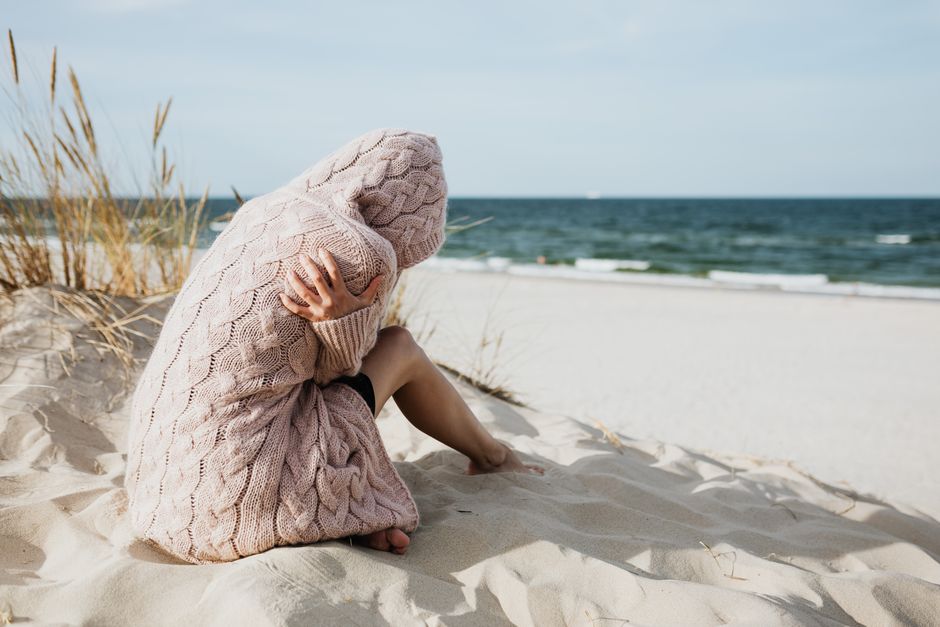Pessoa sentada na areia de uma praia, cobrindo o rosto com um cobertor de tricô, transmitindo uma sensação de reflexão ou tristeza em um ambiente costeiro ao ar livre.
