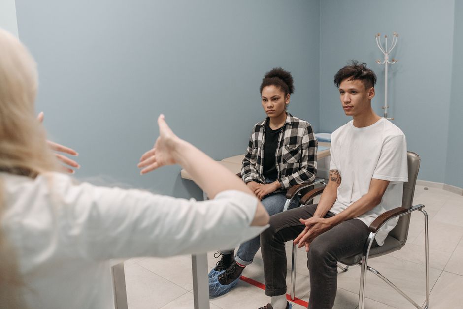 Duas pessoas participando de uma sessão de terapia ou aconselhamento, com uma profissional conversando com um casal jovem em um ambiente clínico.