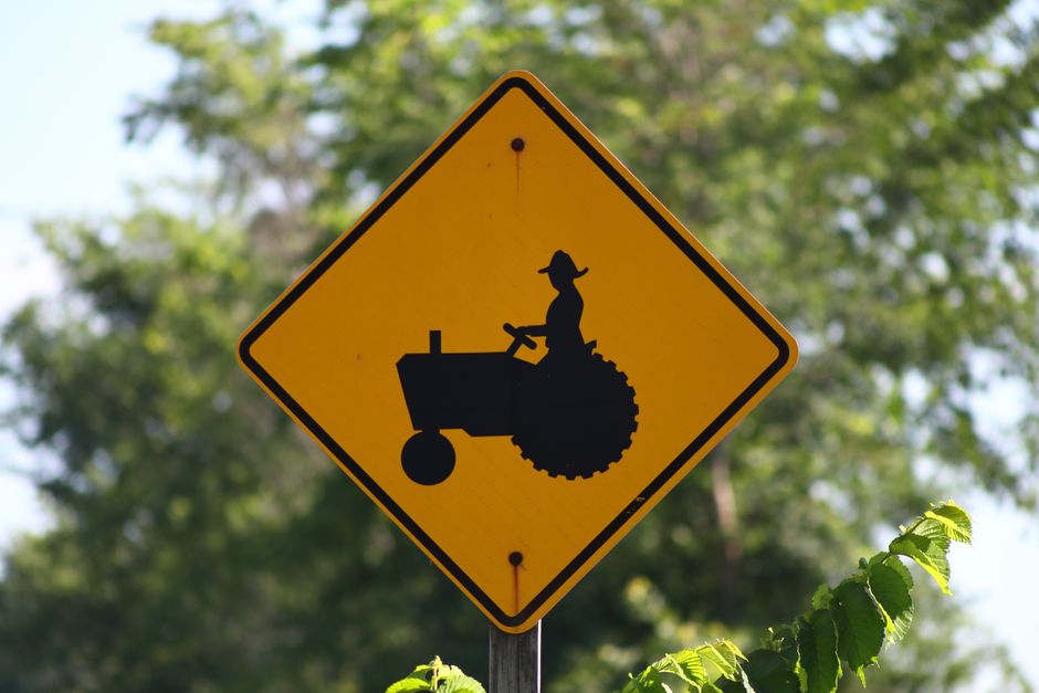 Placa de trânsito com símbolo de uma pessoa dirigindo um trator, indicando área agrícola ou de uso de máquinas agrícolas.