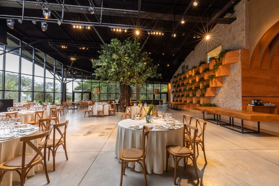 Sala de eventos elegante com iluminação acolhedora, jardim central com árvore decorativa e mesas bem arrumadas para celebrações ou reuniões.