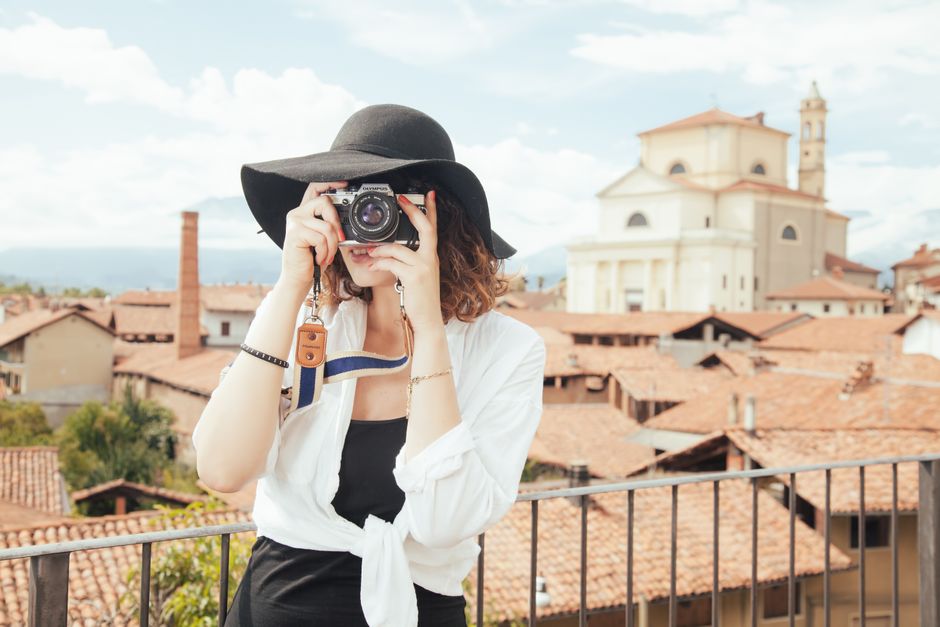 Mulher segurando câmera fotográfica em rua com telhados de barro e igreja ao fundo, em um cenário urbano, para ilustrar temas de fotografia, turismo ou lazer.