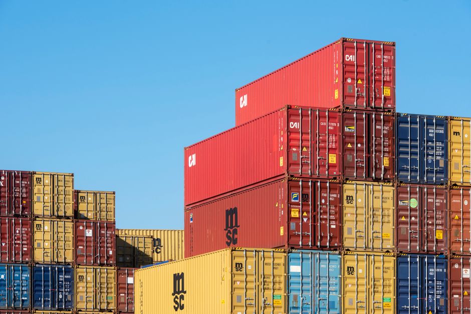 Imagem de contêineres de transporte empilhados em um porto ao céu aberto, representando logística e comércio internacional.