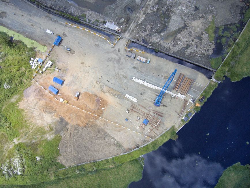 Imagem aérea de um canteiro de obras em fase de construção, com máquinas, materiais e estrutura em desenvolvimento, ao lado de um corpo d'água.