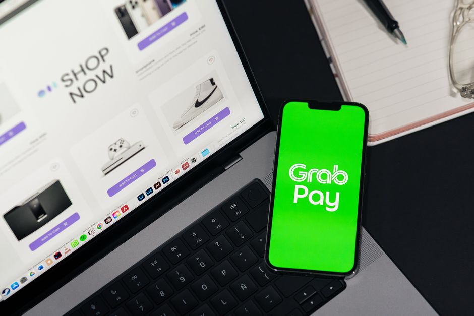 Imagem de um ambiente de trabalho com um laptop exibindo uma loja virtual e um smartphone com o aplicativo Grab Pay, indicando integração de pagamento digital e comércio eletrônico.