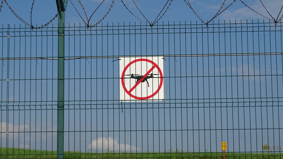 Imagem de uma cerca de segurança com um aviso de proibição de drones, simbolizando restrições ou regulamentos de voo.