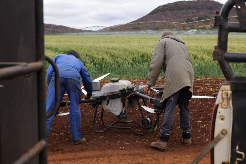 Dois agricultores preparando um drone agrícola para aplicação de insumos em um campo de lavoura com paisagem de morros ao fundo.