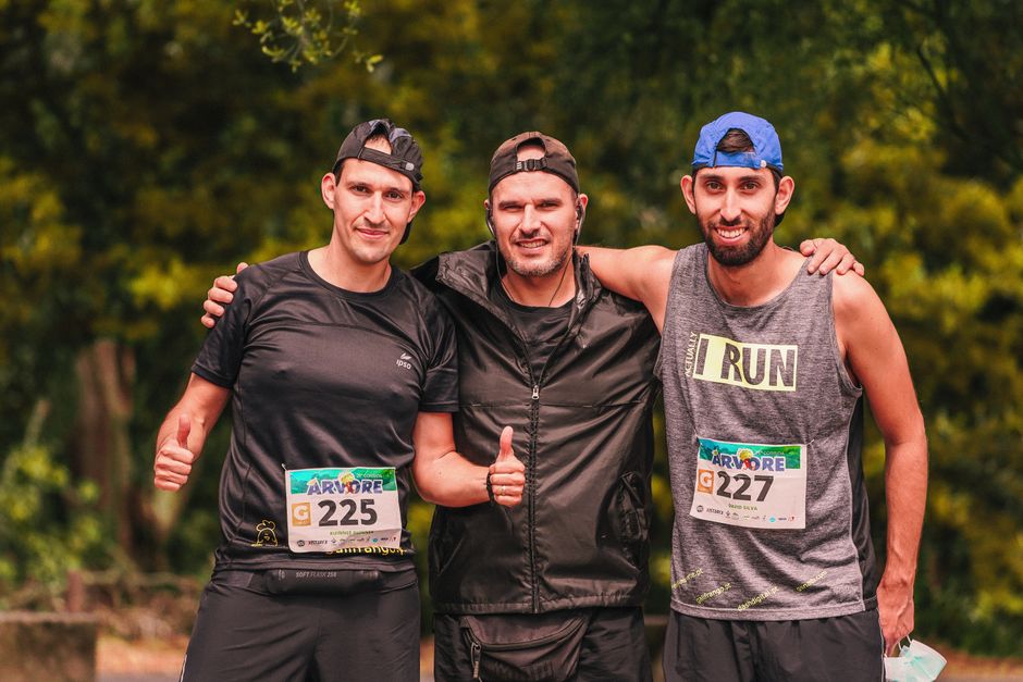 Três homens alfinetados sorrindo após uma corrida ou evento esportivo ao ar livre, celebrando a conquista.