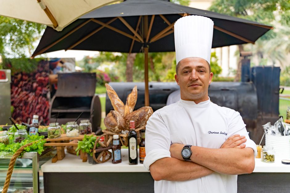 Chef profissional posando com braços cruzados em feira ao ar livre, destacando-se por sua postura de confiança em um evento de gastronomia.