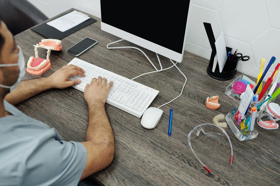 Profissional de saúde utilizando computador em consultório odontológico com modelos de arcadas dentárias ao redor, em um ambiente de atendimento ao paciente.