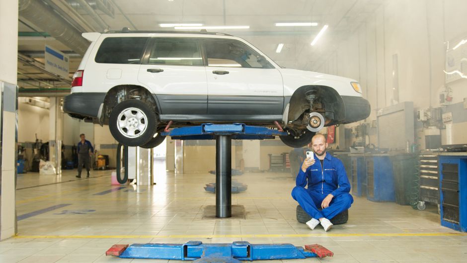 Homem posando com um carro elevado em um ambiente de oficina mecânica, possivelmente para um post sobre manutenção automotiva ou dicas de reparos.