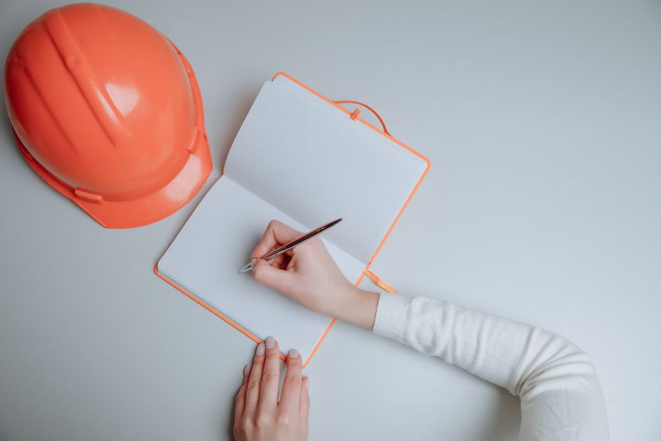 Pessoa anotando informações em um caderno ao lado de um capacete de segurança, simbolizando planejamento ou projetos relacionados à segurança no trabalho ou engenharia.