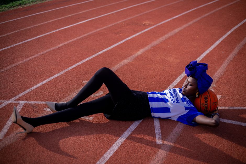 Mulher de traje esportivo de basquete, descansando na pista de corrida, apoiando a cabeça no formato de bola de basquete, simbolizando cansaço e dedicação no esporte.