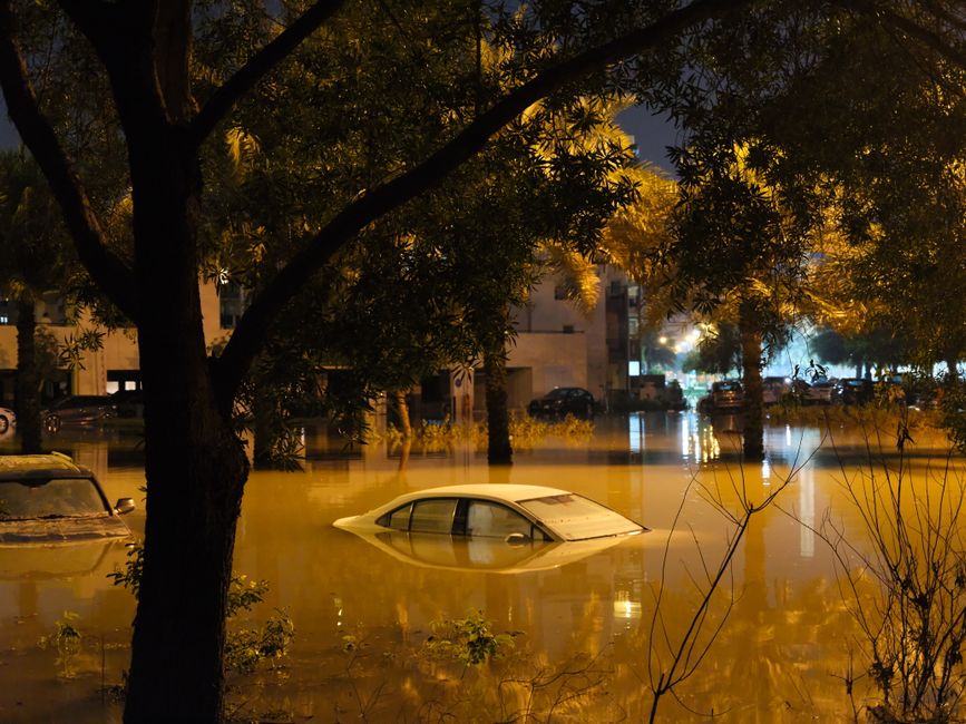 Imagem de uma rua residencial alagada à noite, com um carro parcialmente submerso na água, destacando os efeitos de enchentes urbanas.