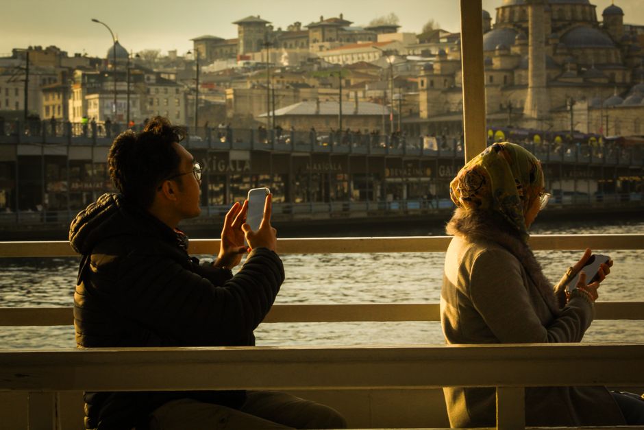 Duas pessoas sentadas em um banco de igreja, usando seus smartphones e apreciando uma vista urbanística ao pôr do sol, em um espaço público às margens de um rio.