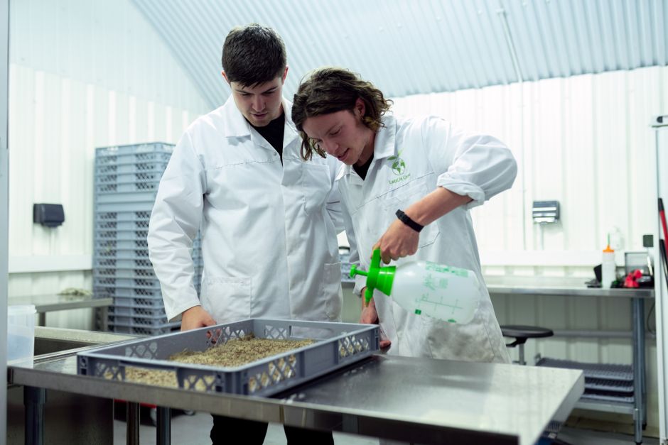 Dois pesquisadores em laboratório analisando uma substância ou produto em um ambiente de pesquisa científica com equipamentos industriais ao fundo.