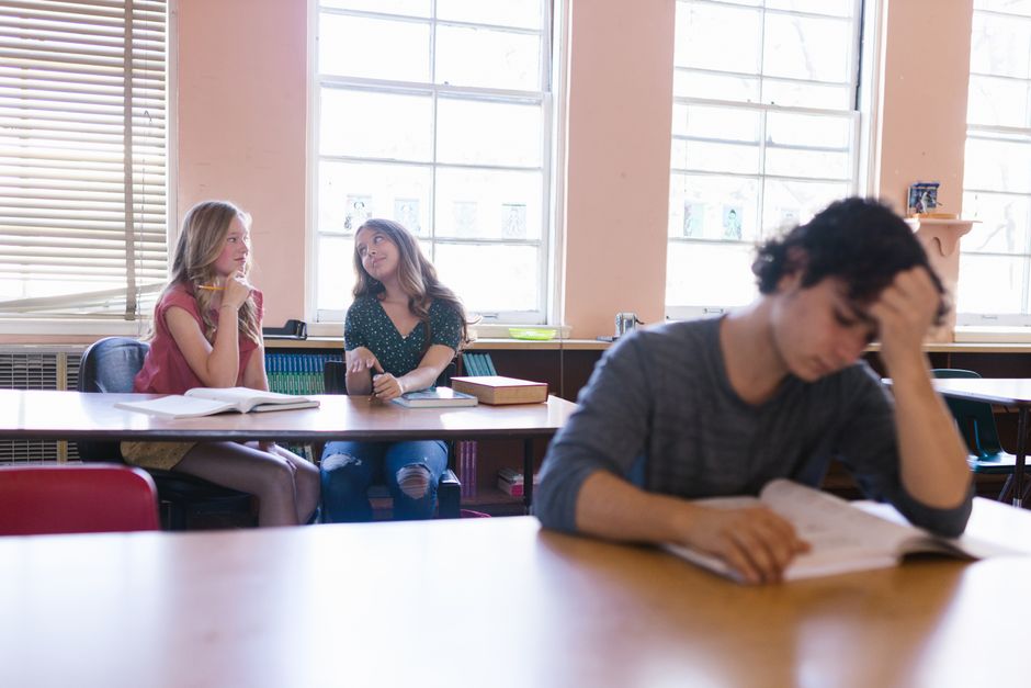 Estudantes em sala de aula com um aluno aparentando cansaço ou frustração enquanto duas colegas conversam ao fundo, refletindo sobre dificuldades acadêmicas.