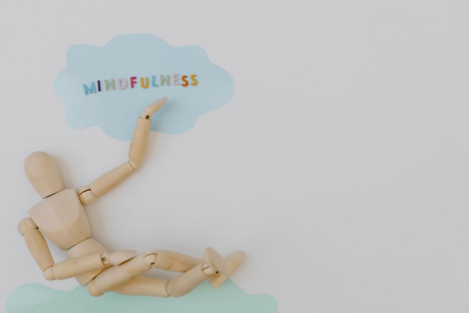 Ilustração com um manequim de madeira deitado e apontando para uma nuvem com a palavra 'Mindfulness', simbolizando prática de atenção plena e bem-estar mental.